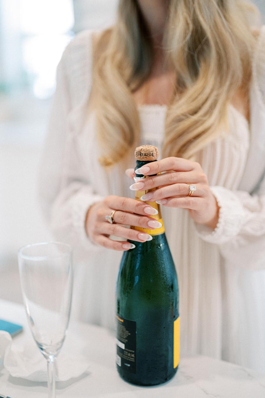 Person in white dress opening a champagne bottle, with a champagne flute on a marble countertop, ready for a celebration.