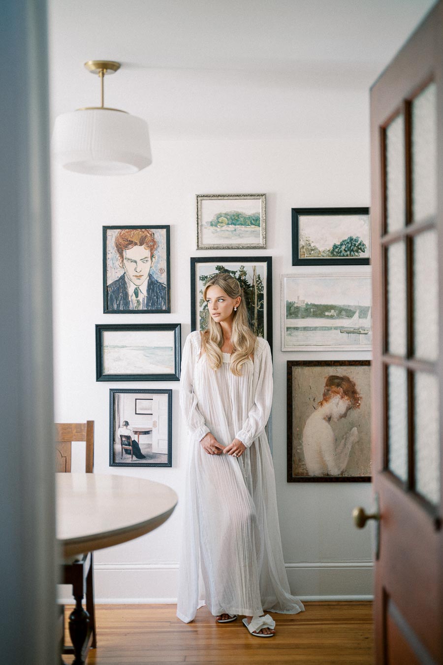 A woman in a flowing white dress stands in a bright, airy room with wooden flooring, surrounded by a gallery wall of eclectic paintings and artwork in various styles and frames.