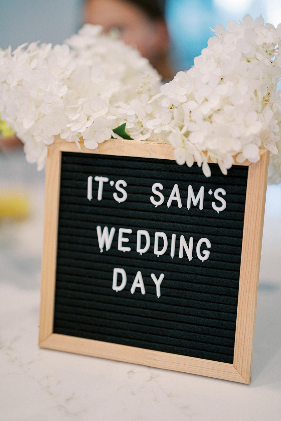 A decorative sign with white lettering on a black background reading It's Sam's Wedding Day, framed by white flowers, used as a wedding reception decoration.