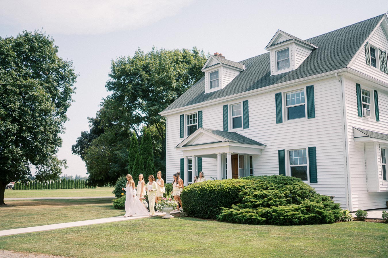 A group of bridesmaids in pastel dresses walking along a pathway in front of a large white colonial-style house with green shutters, surrounded by lush greenery and trees on a sunny day.