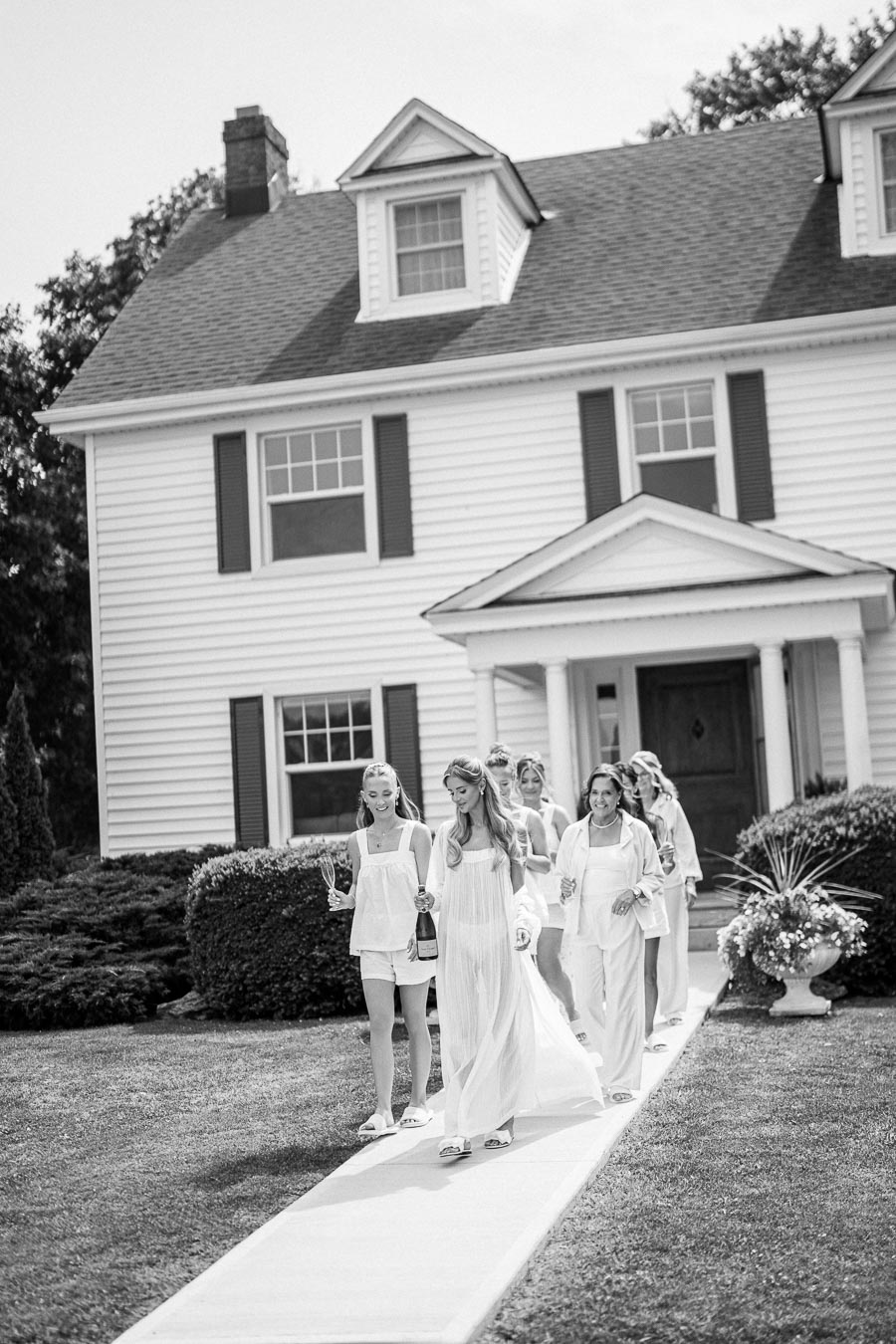 Bridal party walking down path from charming white house, women in light dresses holding champagne and glasses, outdoor celebration.