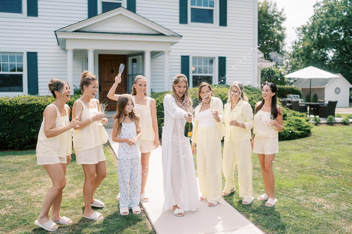 Group of women celebrating outdoors with champagne, wearing matching pastel outfits, in front of a white house.