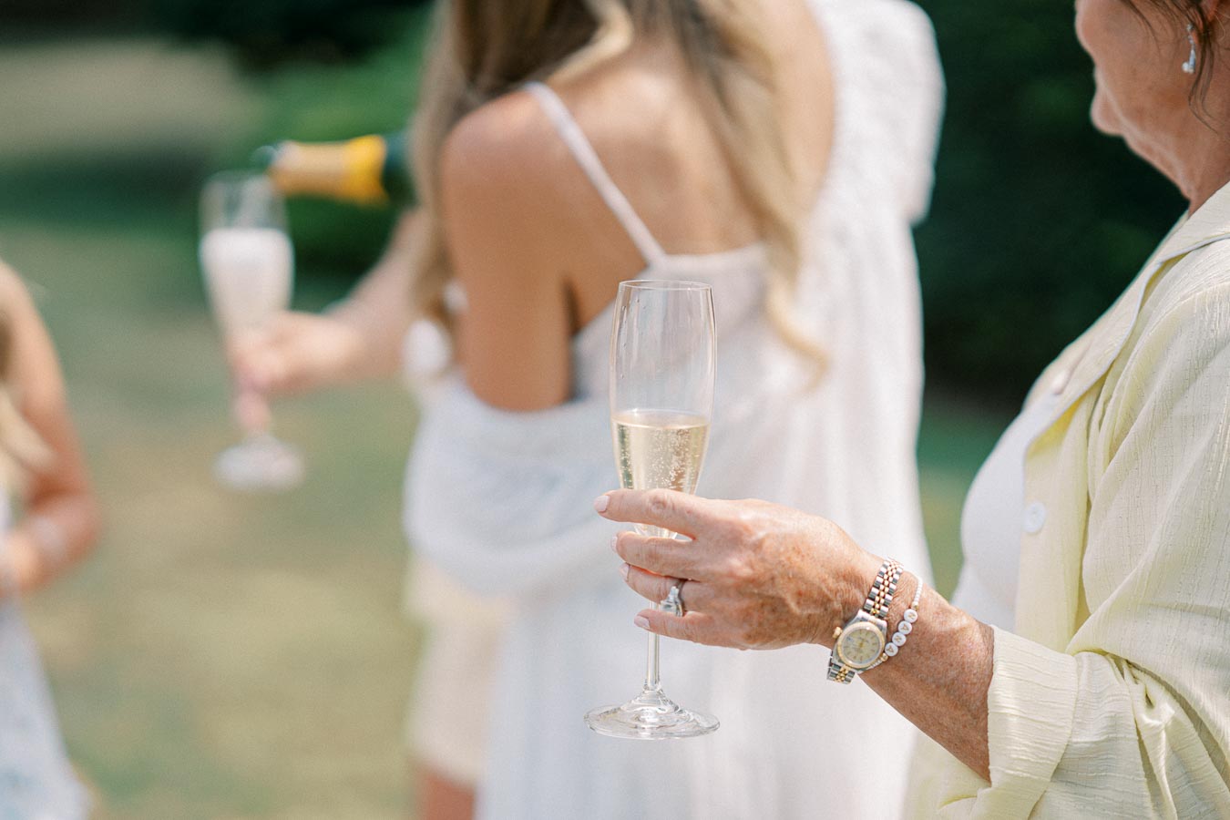 Group of women celebrating outdoors with champagne flutes, focus on hands holding glasses and pouring bubbly in elegant setting.