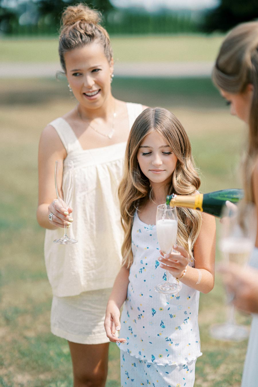 Two women and a young girl enjoying a sunny outdoor picnic while holding champagne glasses, with greenery in the background.