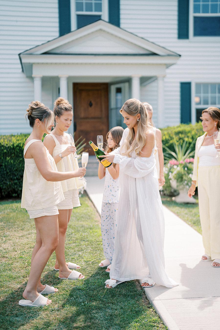 A group of women celebrating outdoors with champagne in a garden setting, wearing matching comfortable white and pastel outfits, in front of a classic white house with blue shutters.