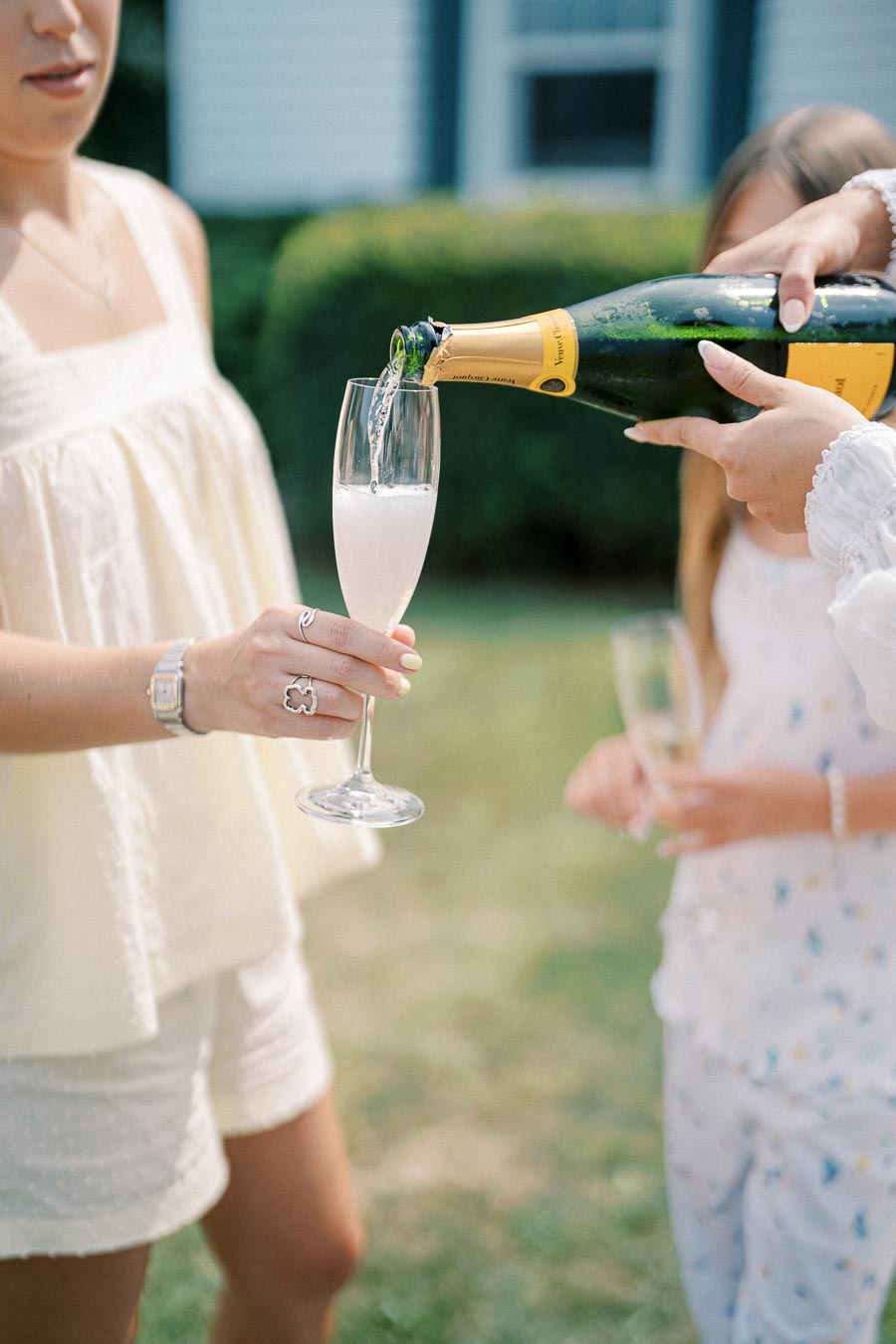 Woman in white summer outfit holding a champagne glass while another person pours champagne outdoors, with a blurred background of greenery and a house.