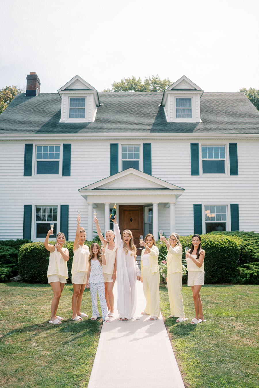 A group of women in matching pastel pajamas and robes celebrating outdoors with champagne in front of a white house with blue shutters, creating a joyful pre-wedding atmosphere.