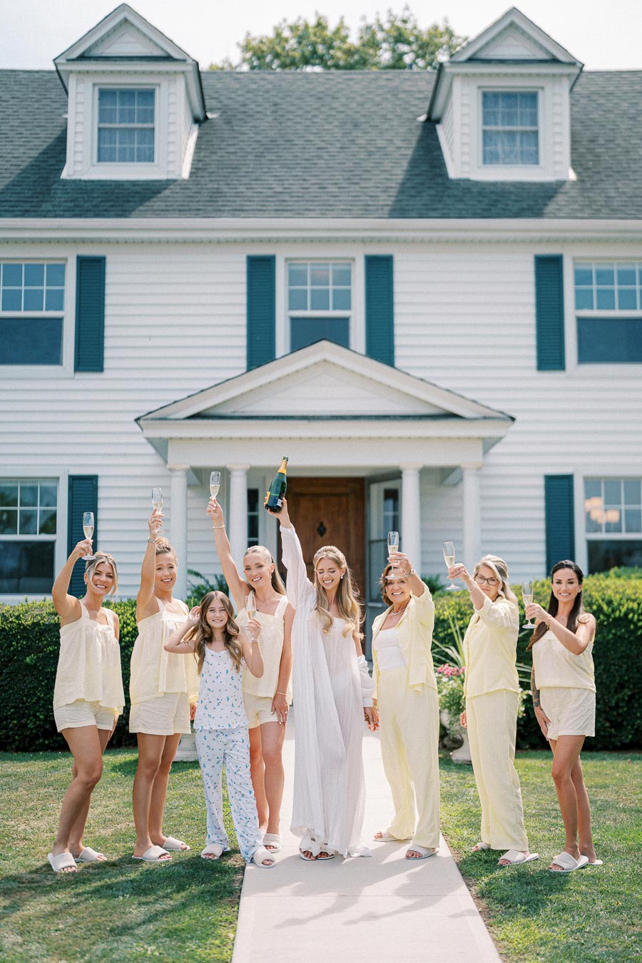A group of women in matching pastel loungewear celebrating with champagne outside a large white house with blue shutters, signifying a joyful gathering or bridal party.
