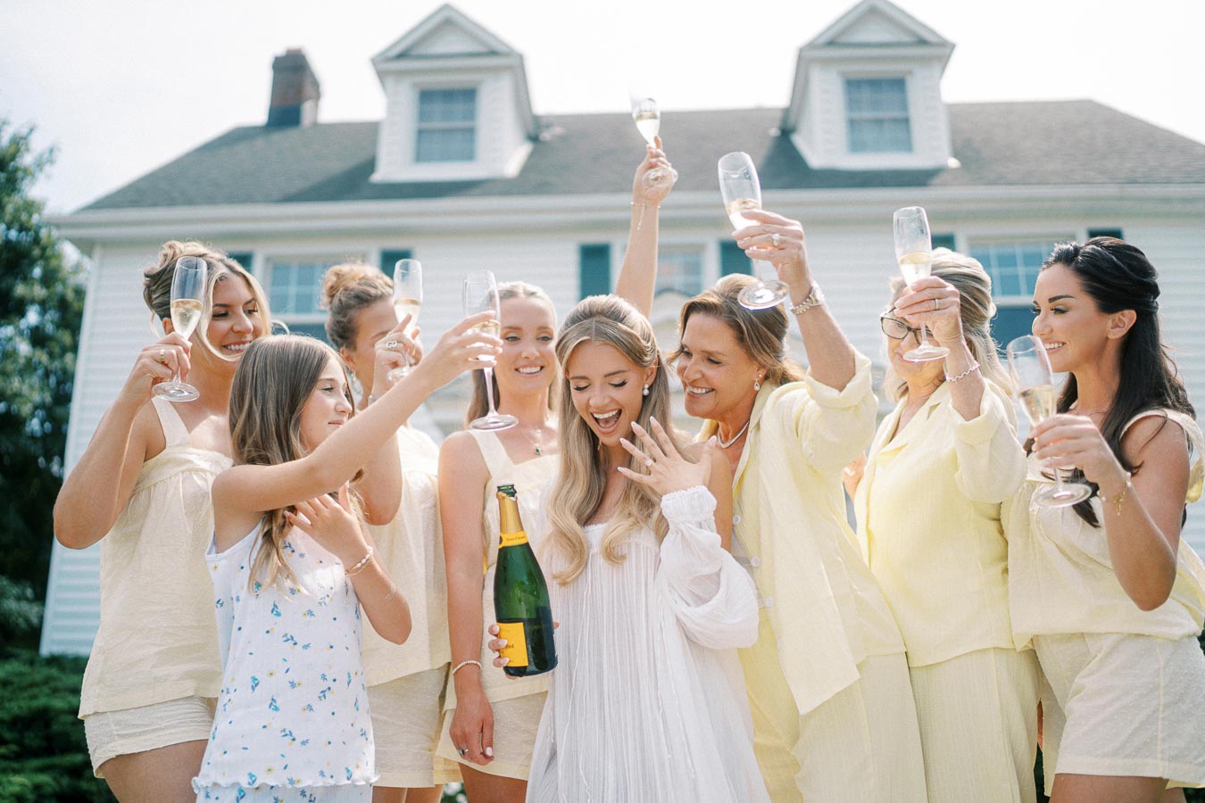 A group of women celebrating outdoors, holding champagne glasses aloft in a joyful toast, with one woman in a white dress holding a champagne bottle, in front of a house under a bright sky.