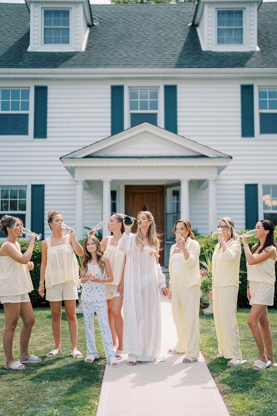 A group of women in loungewear celebrating with champagne outside a white colonial-style house.