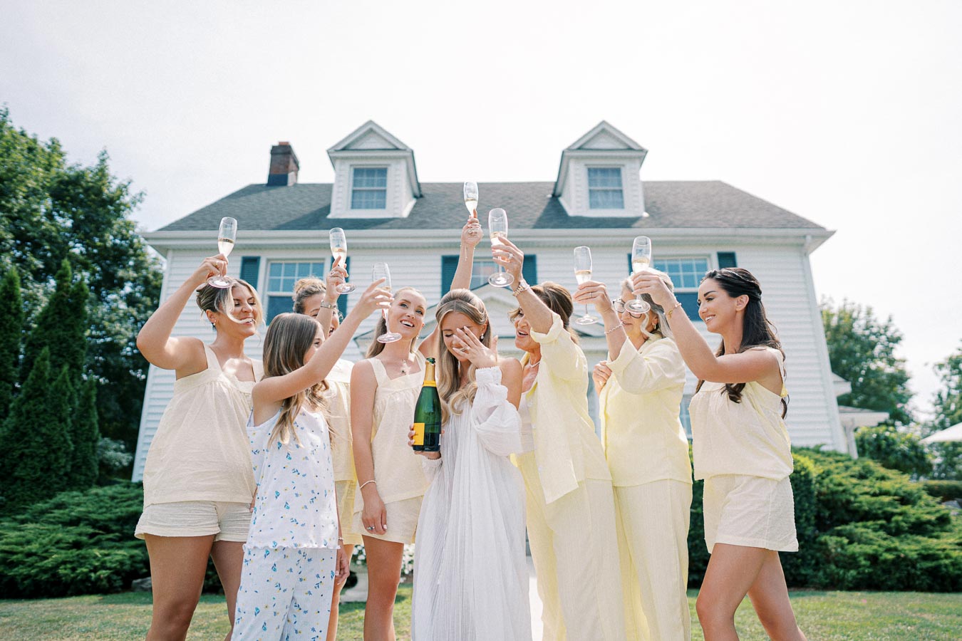 Group of women in matching outfits celebrating with champagne outdoors in front of a house, symbolizing friendship and special occasions.