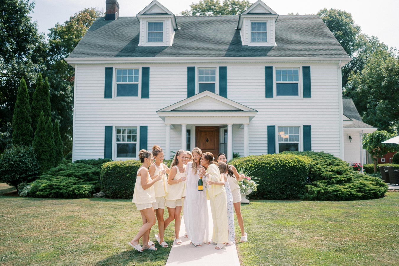 Bridal party celebrating outside a charming white colonial house, with bridesmaids in light yellow outfits and the bride in a white dress holding a champagne bottle.