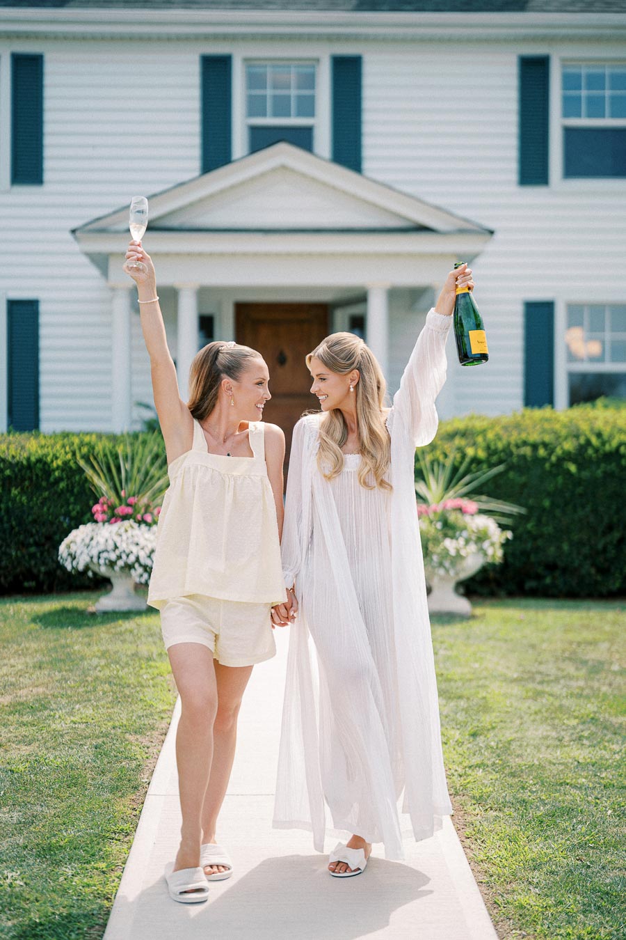 Two women celebrating with champagne, standing on a walkway in front of a white house, holding hands and smiling.