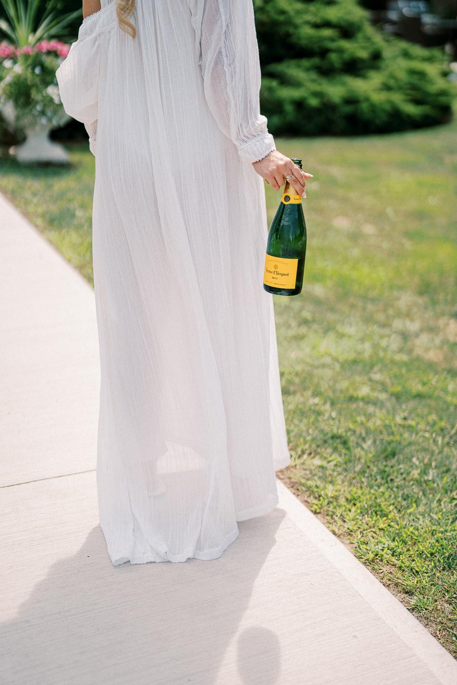 A person in a flowing white dress walking on a sidewalk while holding a bottle of champagne in a green garden setting.