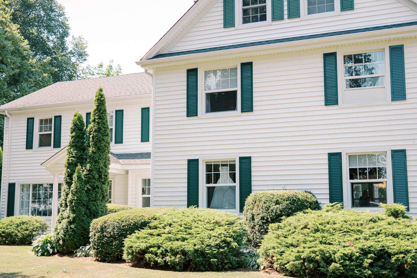 Large white colonial-style house with green shutters and well-manicured shrubs in the front yard.