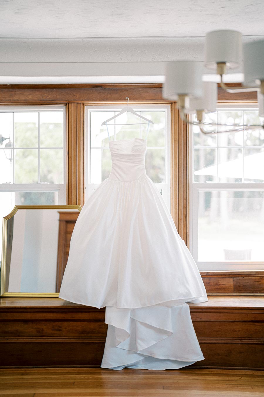 Elegant white wedding dress with full skirt hanging in front of a bright window next to a mirror, emphasizing a classic bridal theme.