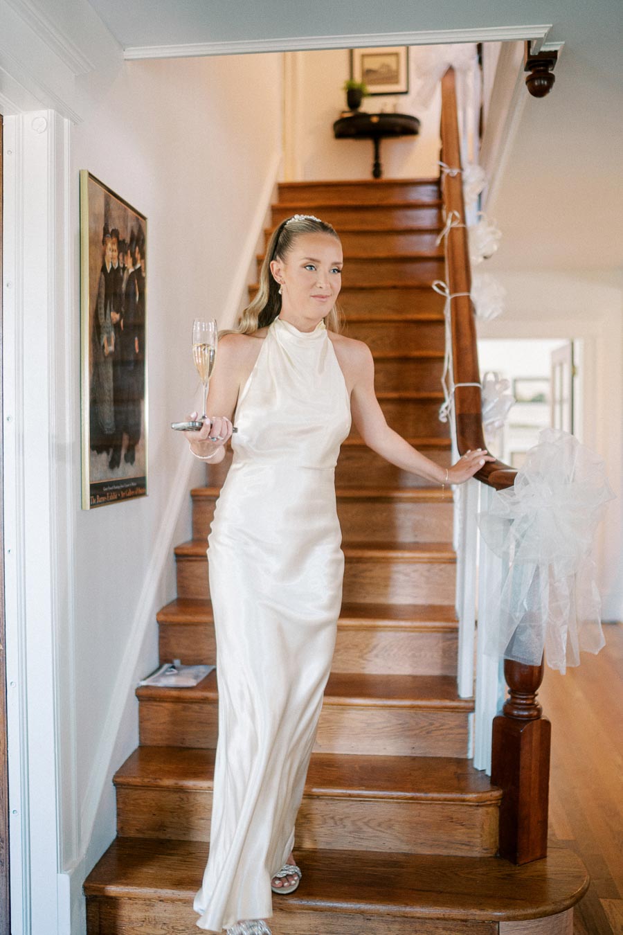 Elegant woman in a white satin gown descends a wooden staircase, holding a champagne flute and a smartphone, with staircase decorated for a special occasion.