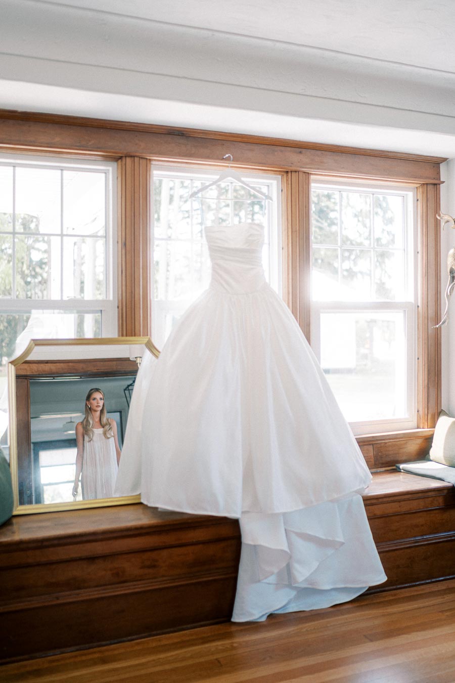 Elegant white wedding dress hanging in front of a large window. The room is filled with natural light, highlighting the flowing layers of the gown. A woman is reflected in a mirror, gazing at the dress.