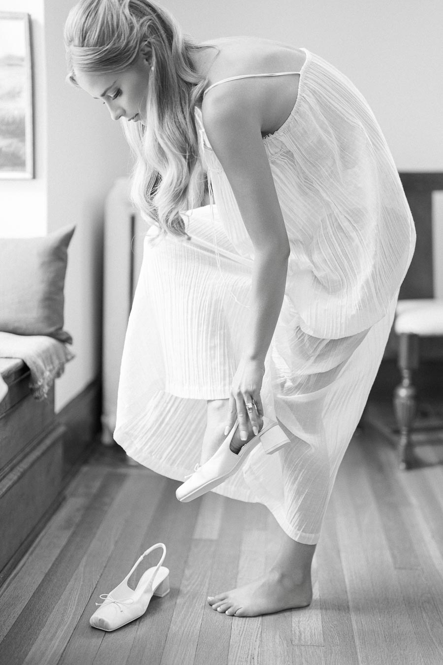 A woman in a flowing white dress gracefully slips on a pair of elegant heels in a softly lit room.