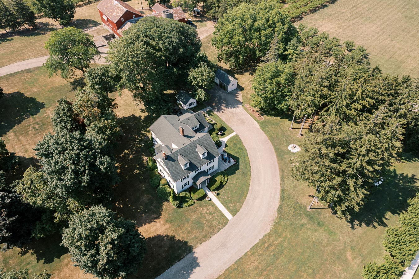 Aerial view of a large country estate with a classic white farmhouse surrounded by lush green trees, a curved driveway, and a red barn in the background.