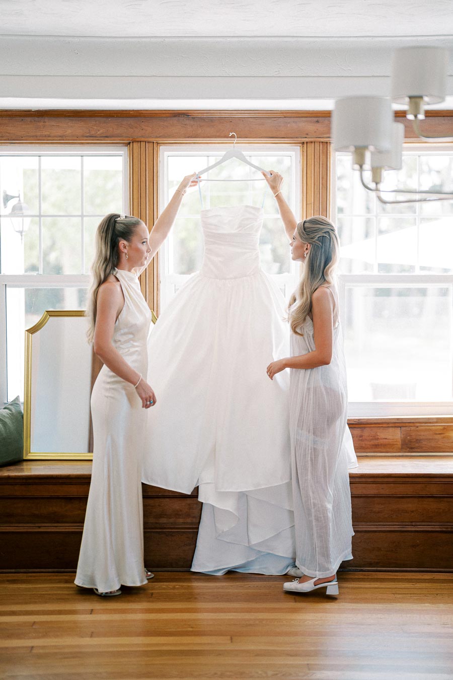 Two women in elegant dresses admire a white wedding gown on a hanger in a sunlit room.