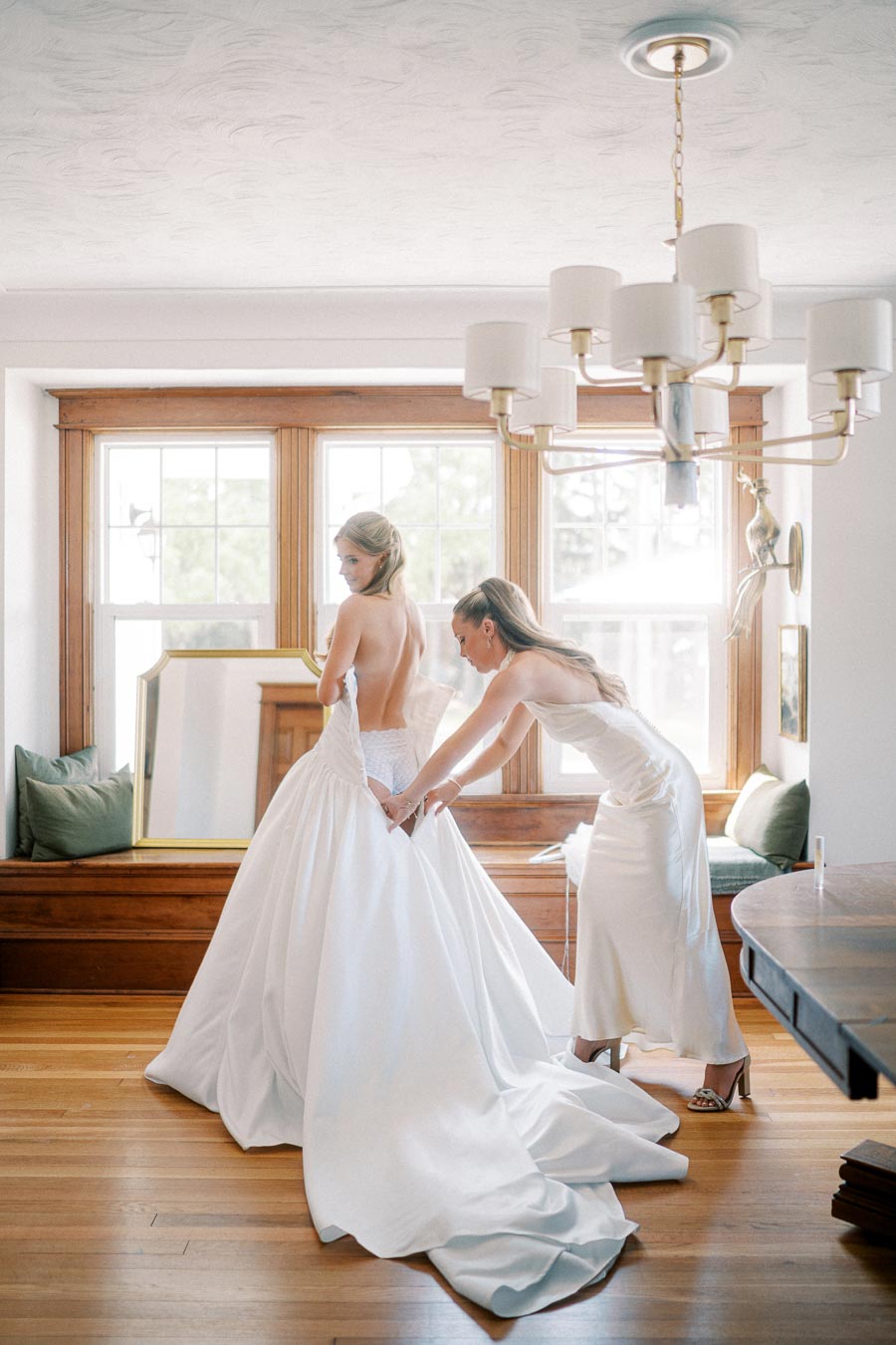 A bride getting dressed in a luxurious white wedding gown with the help of her bridesmaid in an elegantly decorated room with wooden floors and large windows.