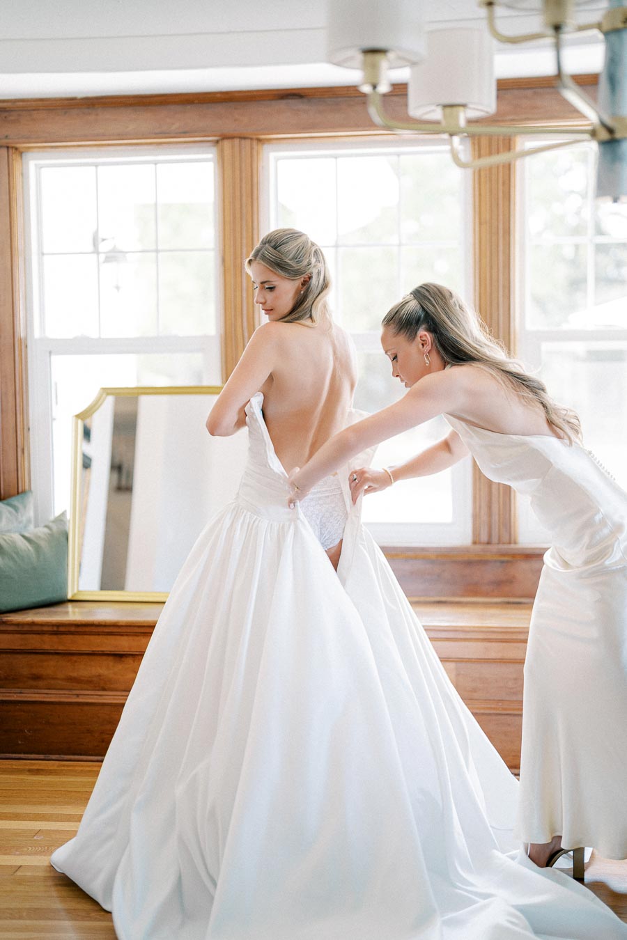Two women in a bright room, one helping the other zip up her elegant white wedding dress, with natural light streaming through large windows.