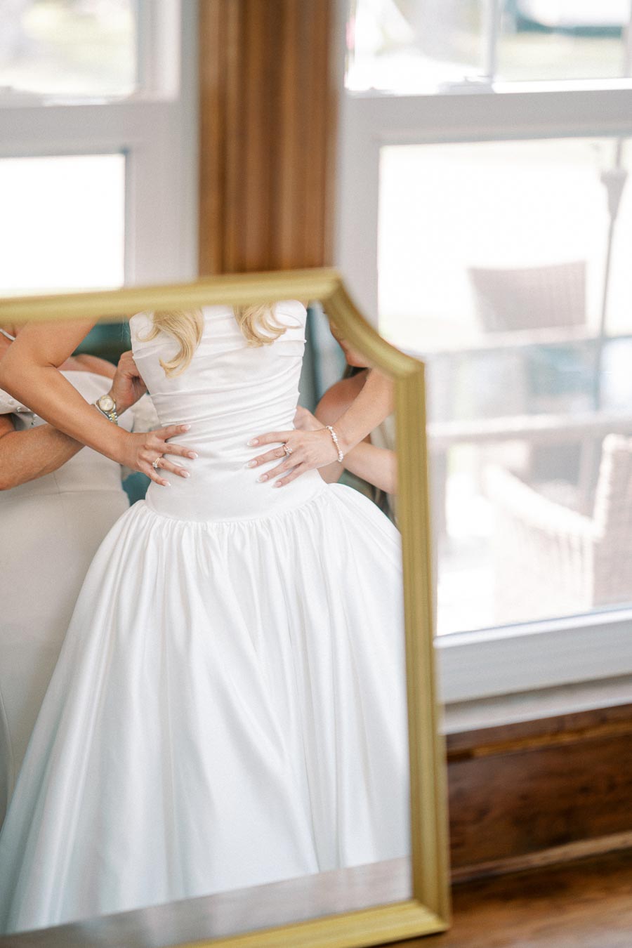 Reflection of a bride in a mirror adjusting her elegant white wedding gown, showcasing intricate design details and a serene preparation moment by a window.