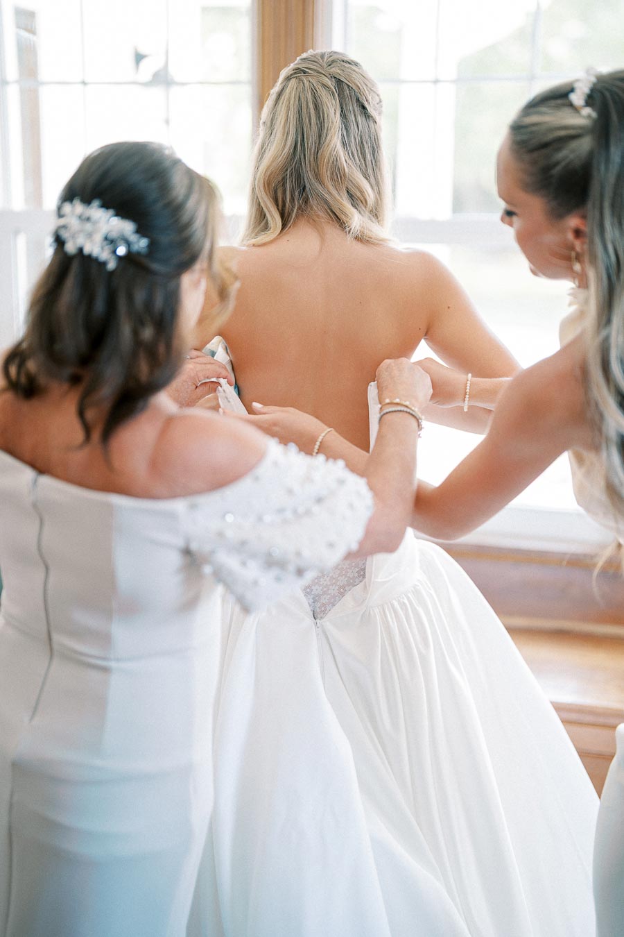 Bridesmaids help a bride get ready by adjusting her elegant white wedding gown near a bright window.