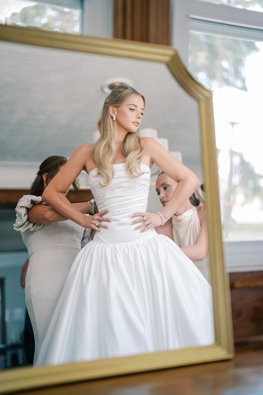 A bride in a white wedding gown is adjusting her dress with the help of her bridesmaids, captured in a large ornate mirror.