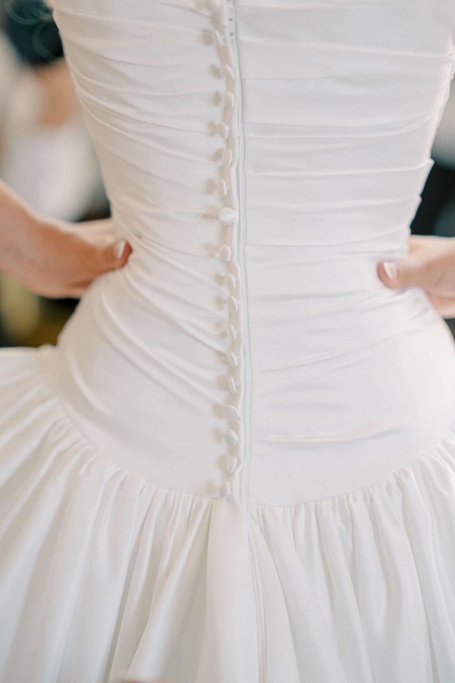 Close-up of a bride wearing a white wedding dress with intricate button detailing on the back, hands on hips.