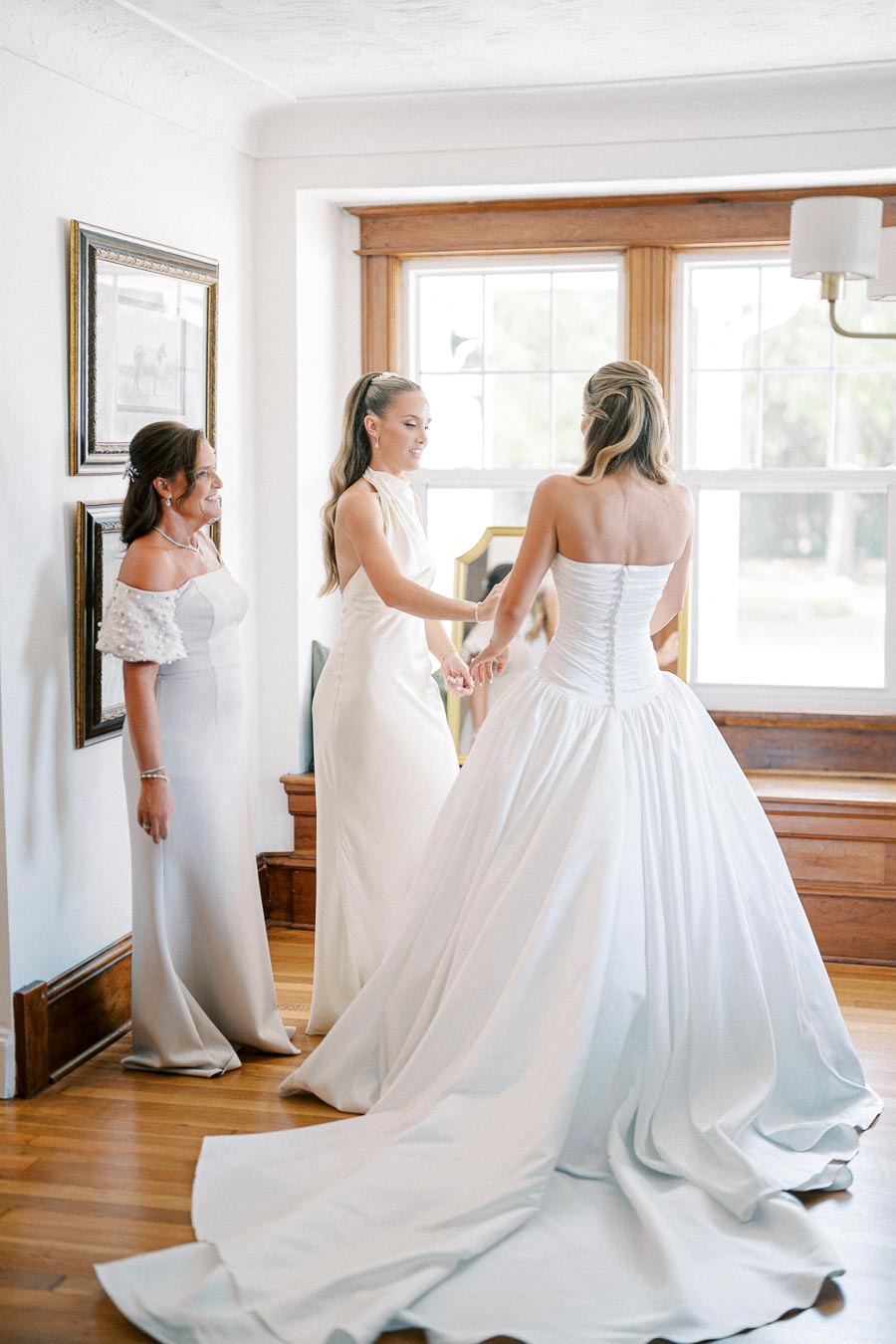 Three women in elegant dresses, with a bride in a classic white gown, preparing in a bright room before a wedding ceremony.