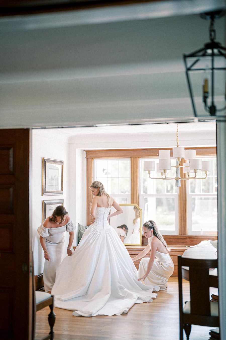 A bride in an elegant white wedding gown is assisted by her bridesmaids in a sunlit room, preparing for her wedding ceremony.