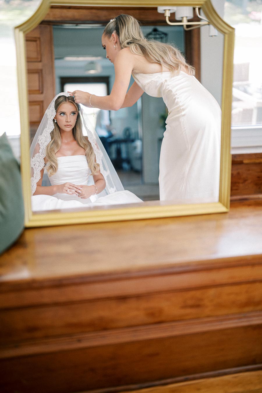 Bride getting ready with her veil adjusted by a bridesmaid, reflected in a mirror, in a serene bridal suite.