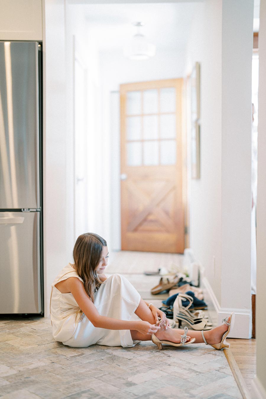 Young woman in a white dress sitting on a tiled floor, adjusting her shoes in a bright hallway.