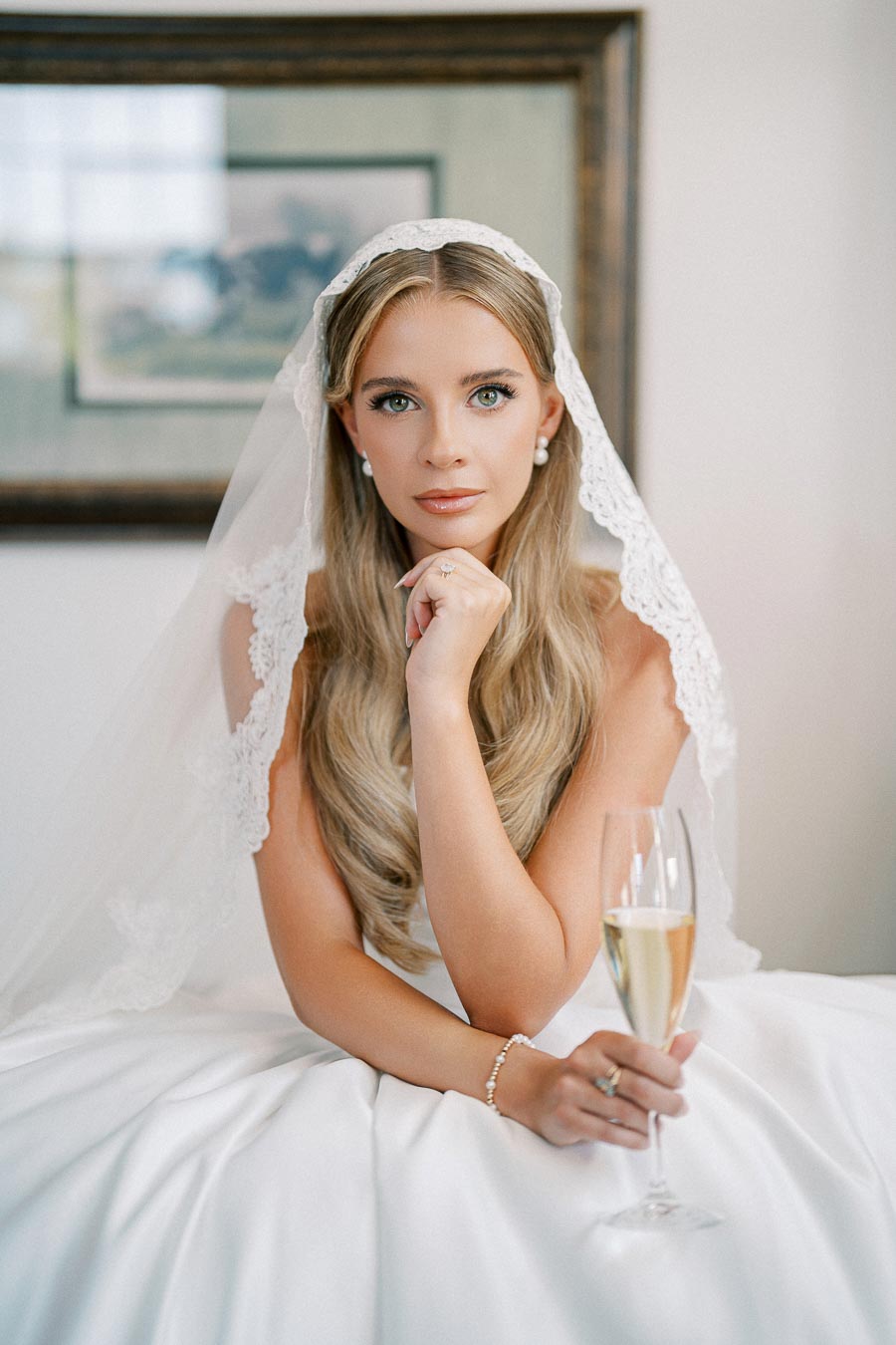 Elegant bride in a white dress and lace veil, holding a champagne glass, sitting pensively with stunning makeup and long wavy hair.