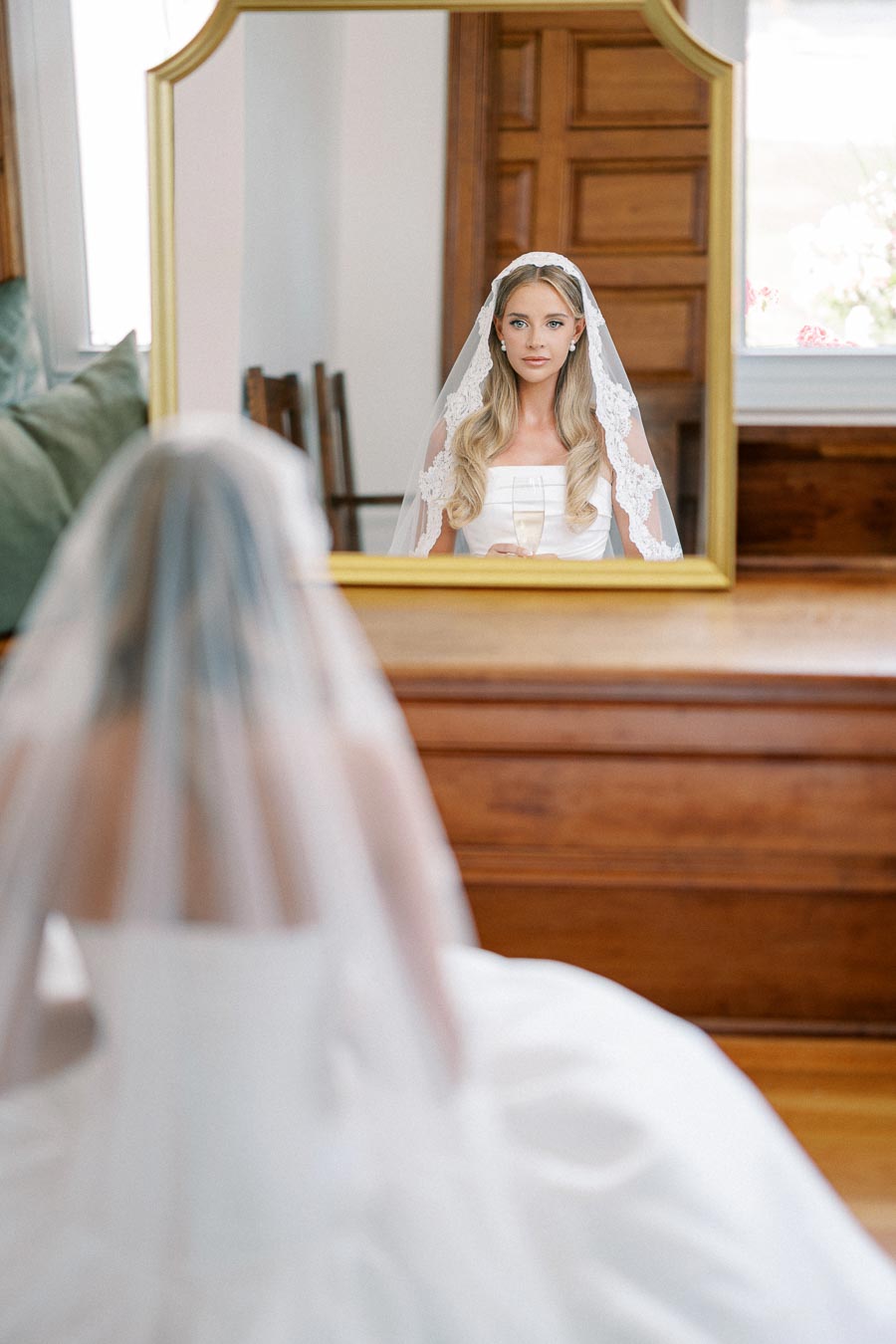 A bride in a white gown and lace veil gazes at her reflection in a large mirror, set in an elegant room with wooden accents.