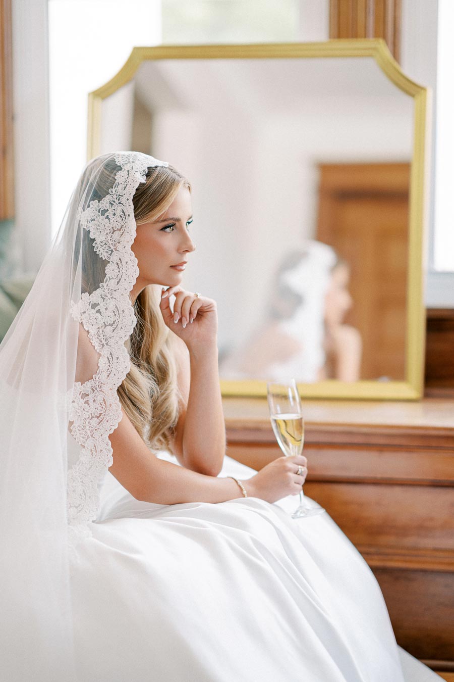 Elegant bride in a white lace veil sitting thoughtfully near a mirror, holding a champagne glass, reflecting bridal beauty and pre-wedding contemplation.