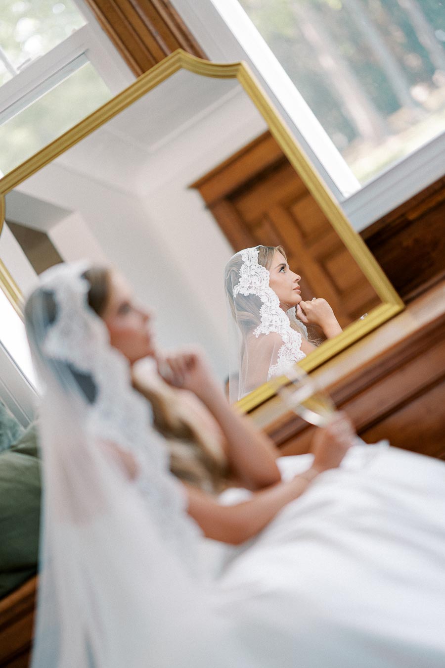 Bridal portrait of a woman in a lace veil, contemplating in front of a mirror while holding a glass of champagne.