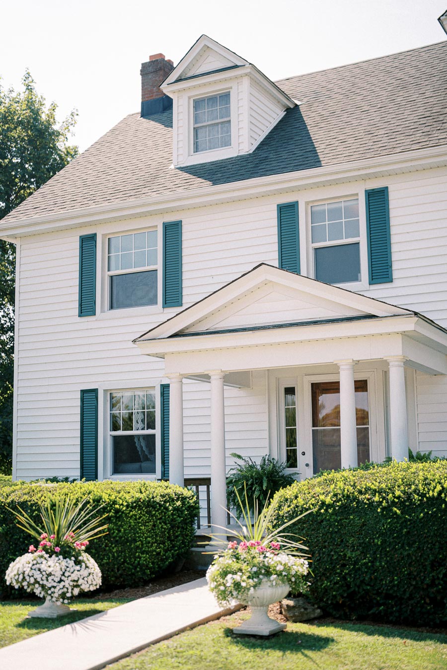 Charming white two-story house with green shutters, a covered front porch, and lush garden landscaping featuring colorful flowers and manicured bushes.