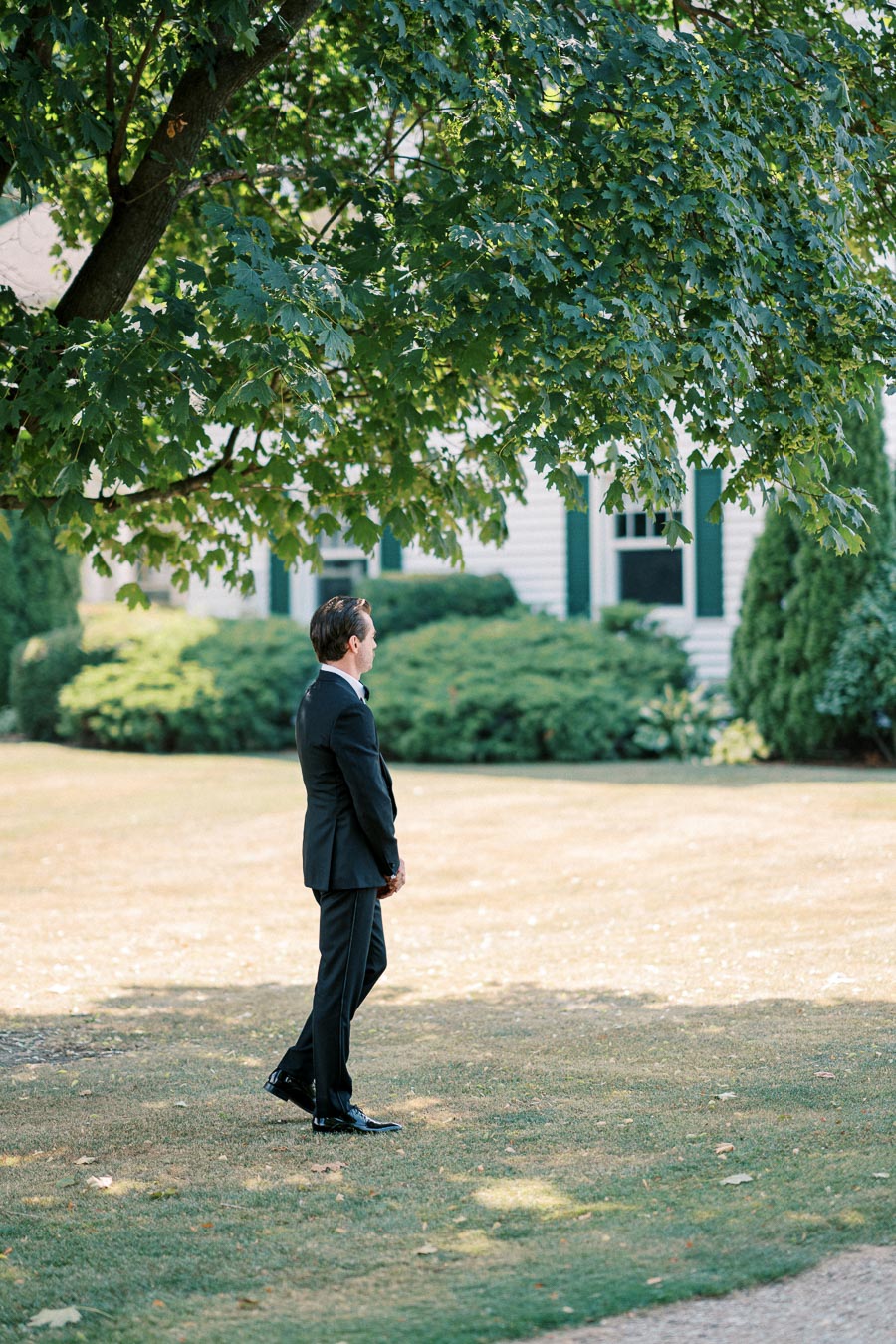 A man in a formal black suit stands on a lawn under the shade of a tree, with a house and greenery in the background, conveying a serene and elegant outdoor setting.