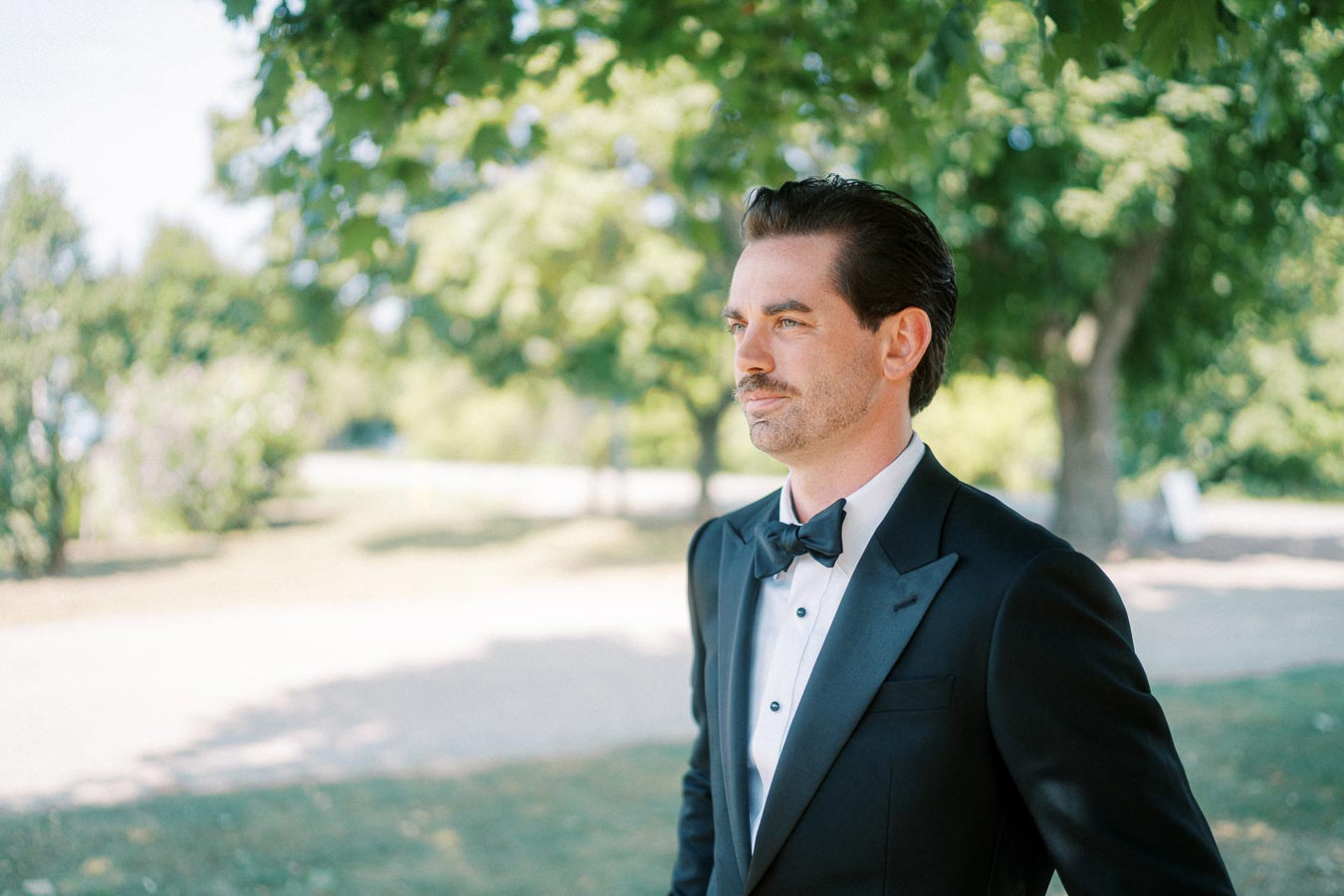 Man in formal black tuxedo with bow tie standing outdoors under tree shade, looking away with a thoughtful expression, park setting in the background.