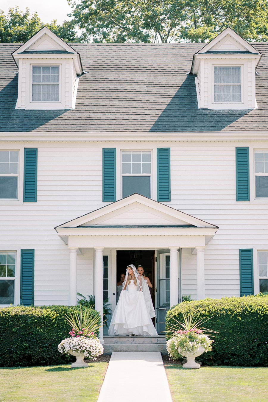 Elegant bride in white dress standing at the entrance of a classic white house with blue shutters, surrounded by lush greenery and flower arrangements, under a clear blue sky.
