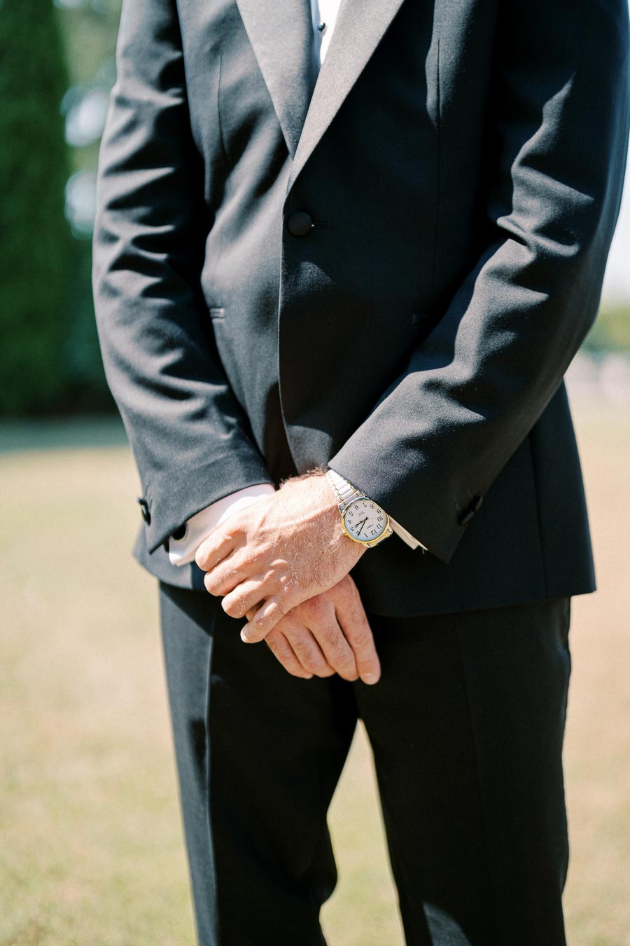 Man in a tailored black suit with a silver watch stands with hands crossed against an outdoor setting.