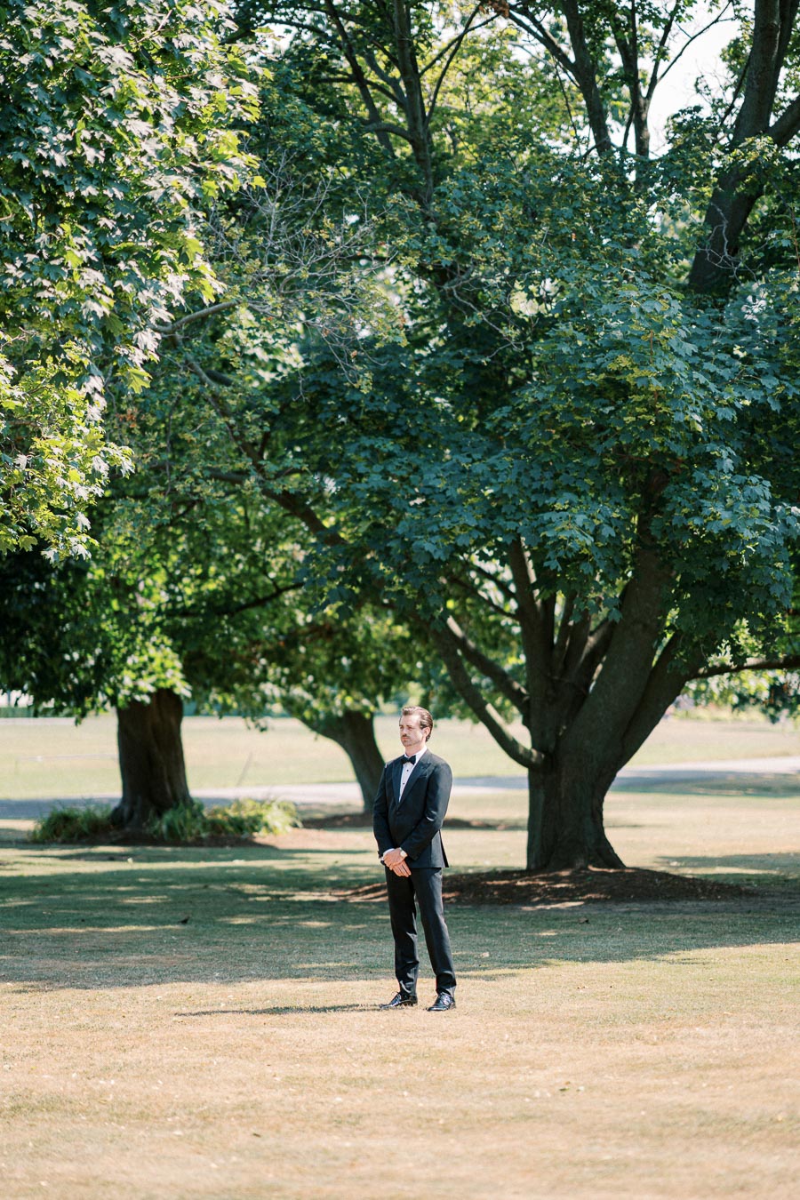 A groom in a black tuxedo stands alone on a sunny day beneath tall green trees in a peaceful park setting.