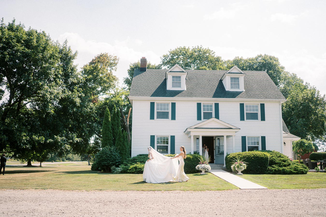 Elegant bride in white gown with bridesmaids outside classic white house on sunny wedding day