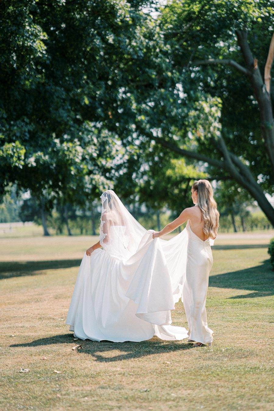 A bride in a flowing white wedding dress walks on a lush green lawn, accompanied by a bridesmaid in a sleek gown, beneath the shade of large trees on a sunny day.