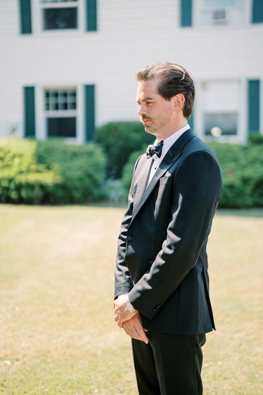 A man in a black tuxedo stands outside in a garden, exuding elegance and sophistication.
