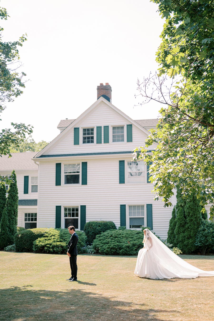 Bride and groom in a garden in front of a large white house, surrounded by greenery, on their wedding day.