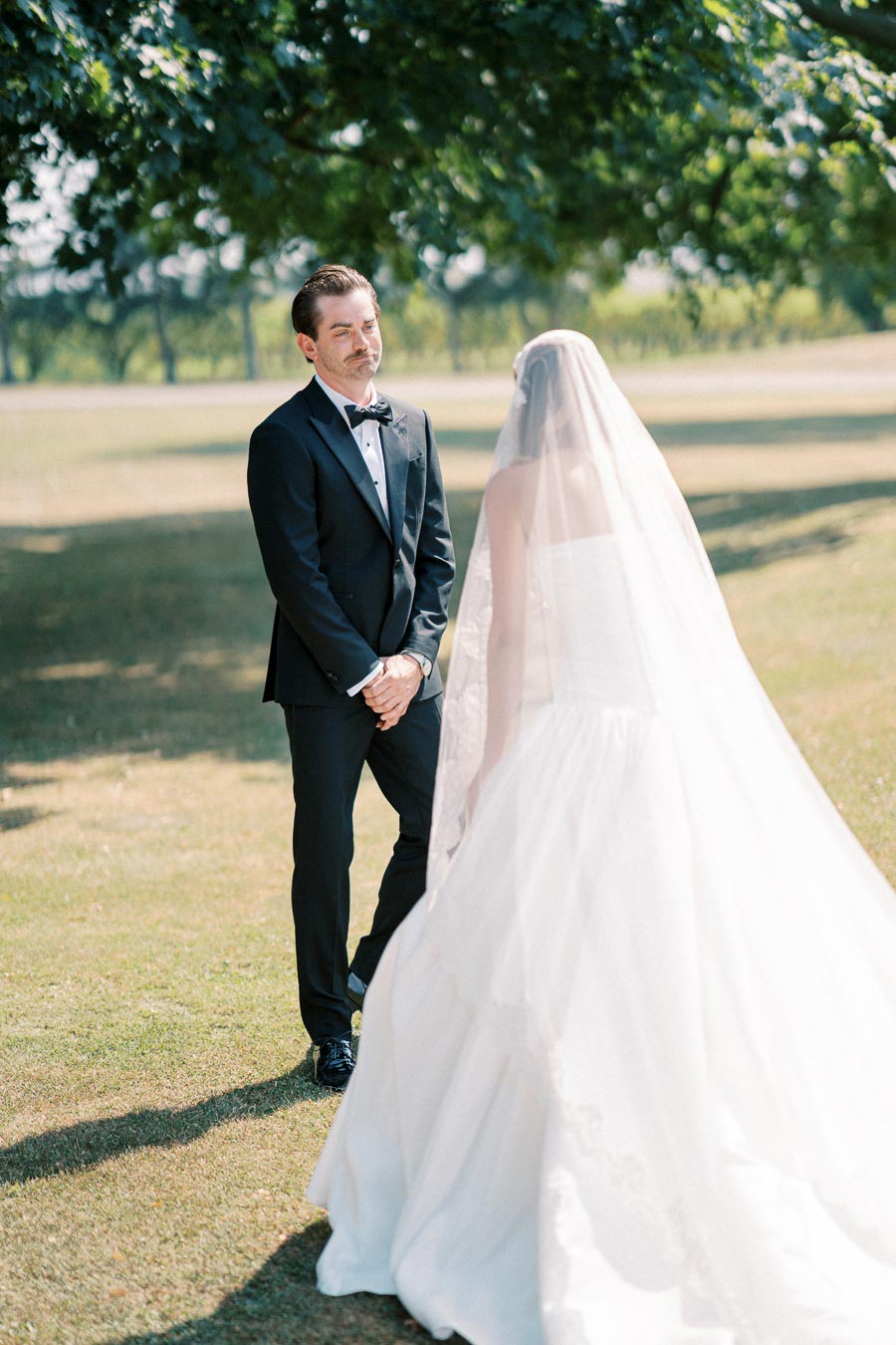 A groom in a black tuxedo and a bride in a white wedding dress share a tender moment outdoors under a tree, creating a romantic wedding atmosphere.